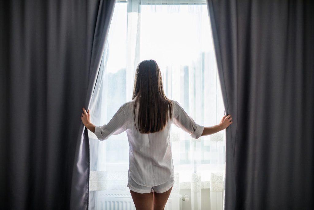 beautiful young woman opening curtains looking through window scaled 1